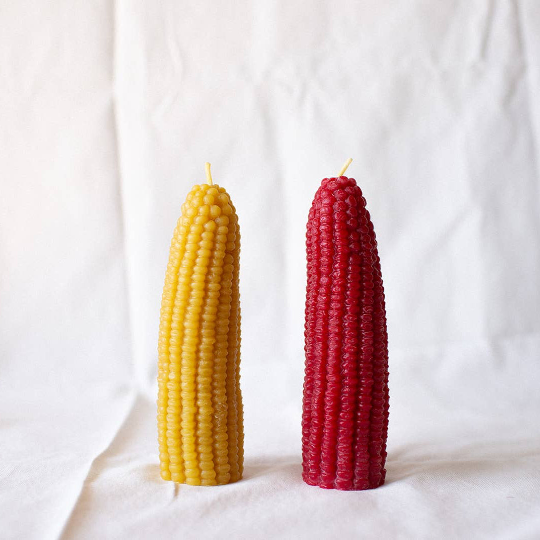 Two corn-shaped candles, one yellow and one red, on a white fabric background.