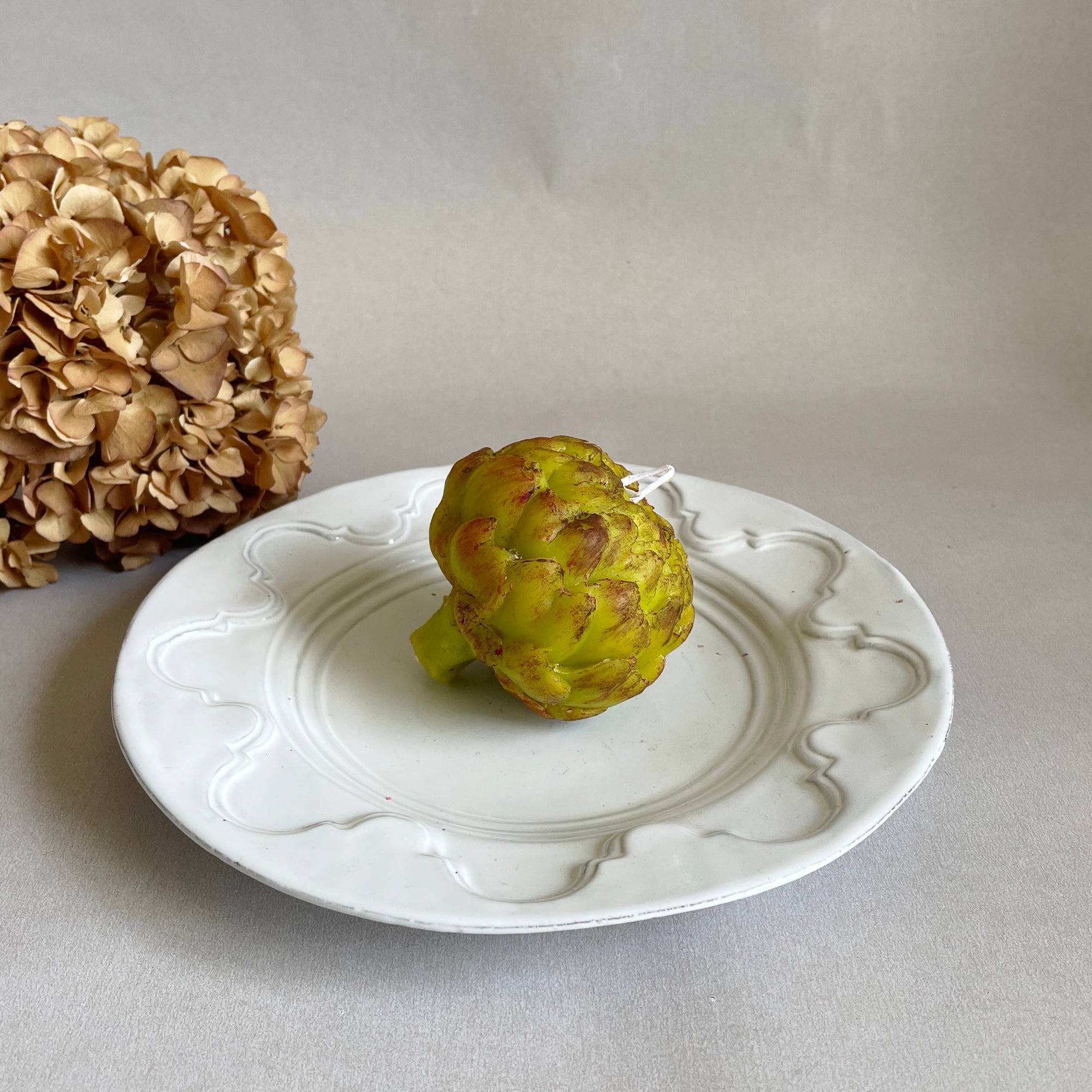 A handcrafted artichoke-shaped candle made from scented bee's wax, displayed on a white plate with a dried flower in the background.