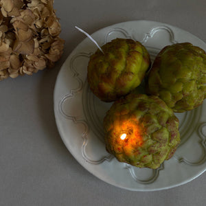 A beeswax scented candle shaped like an artichoke, placed on a white plate, with a real artichoke in the background.