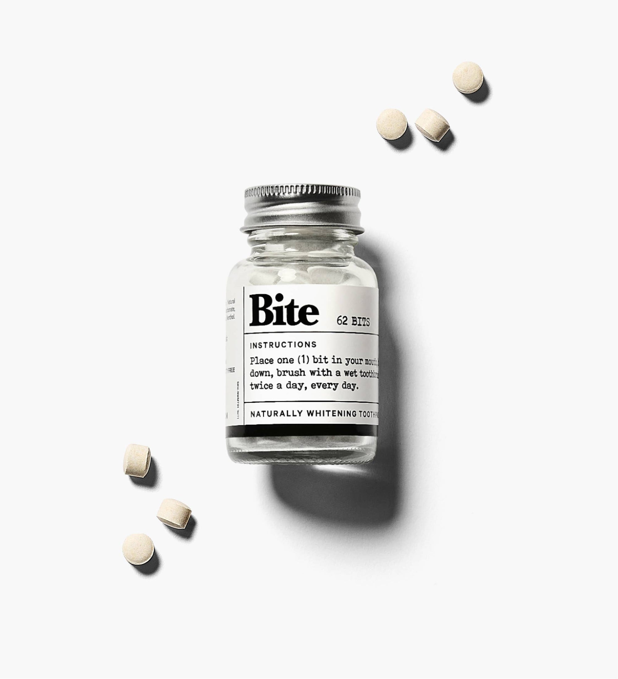 A glass bottle labeled 'bite' containing toothpaste bits, with a few toothpaste bits scattered outside the bottle on a white background.