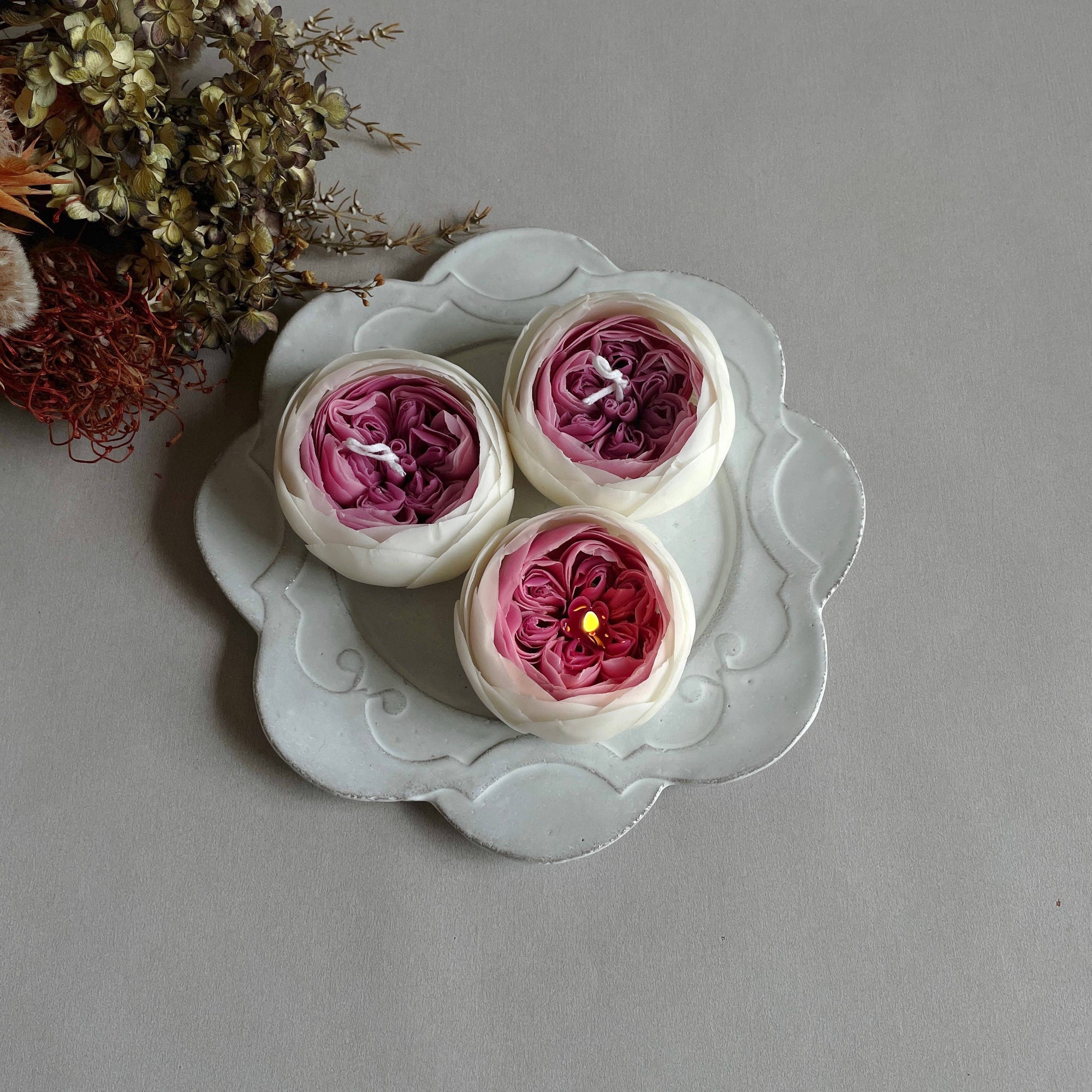 A hand holding a rose-shaped candle with layers of pink and white petals, placed on a white dish with more candles and dried flowers in the background.