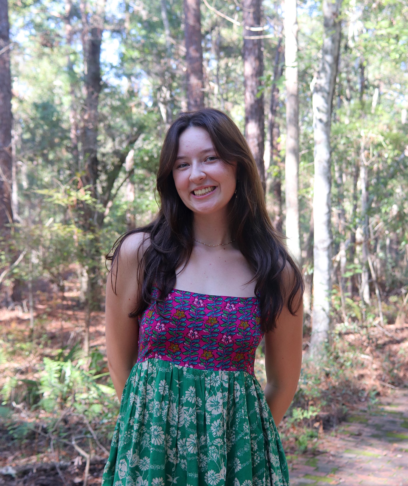 A light skinned woman with long dark hair, wearing a green and purple floral printed dress.