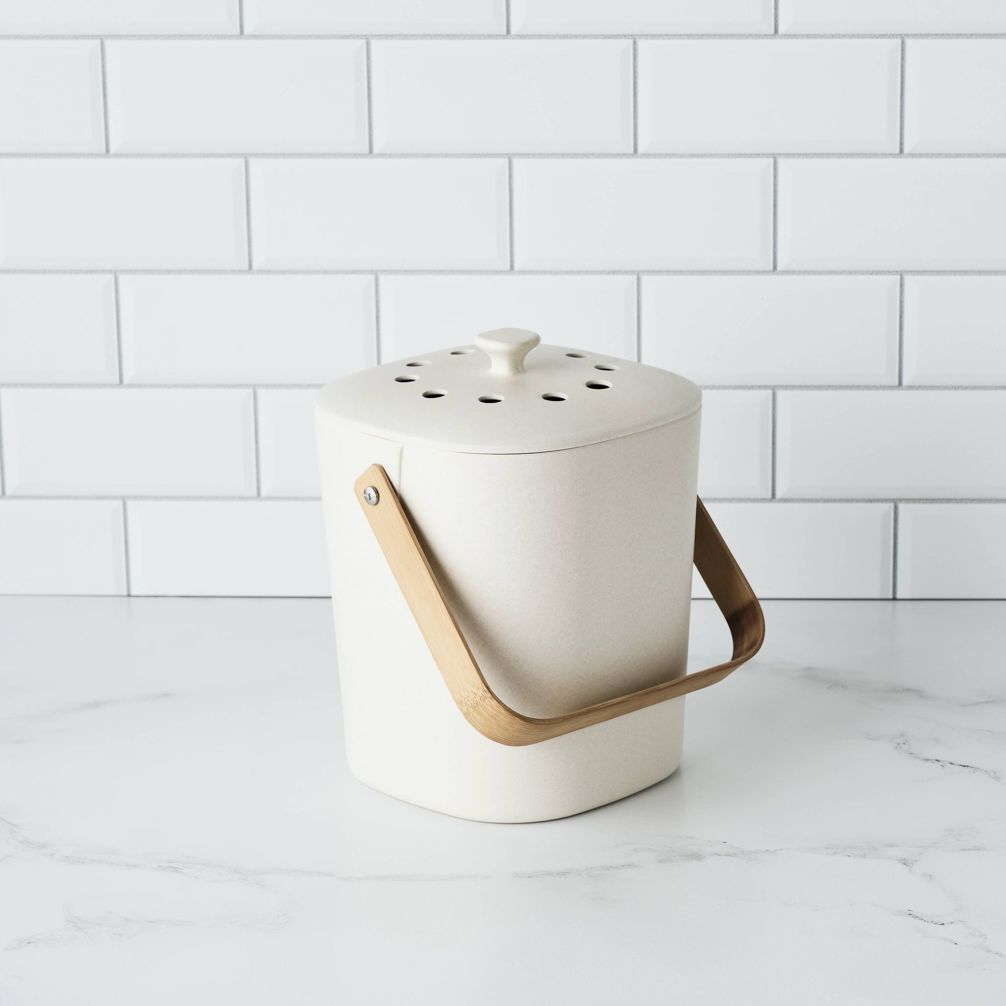 A cream colored composter on a marble counter with a white tile wall behind it. 
