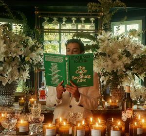 A person holding a "The Guide to Becoming Alive" surrounded by lit candles and flowers. 