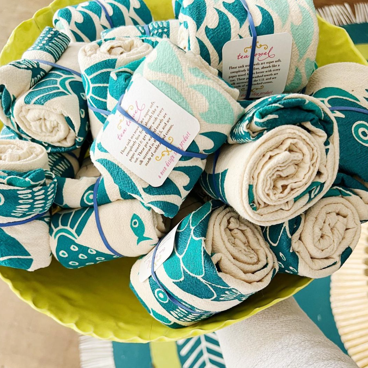 A folded beige cotton tea towel with a silkscreened blue and green wave pattern design, placed on a wooden surface.
