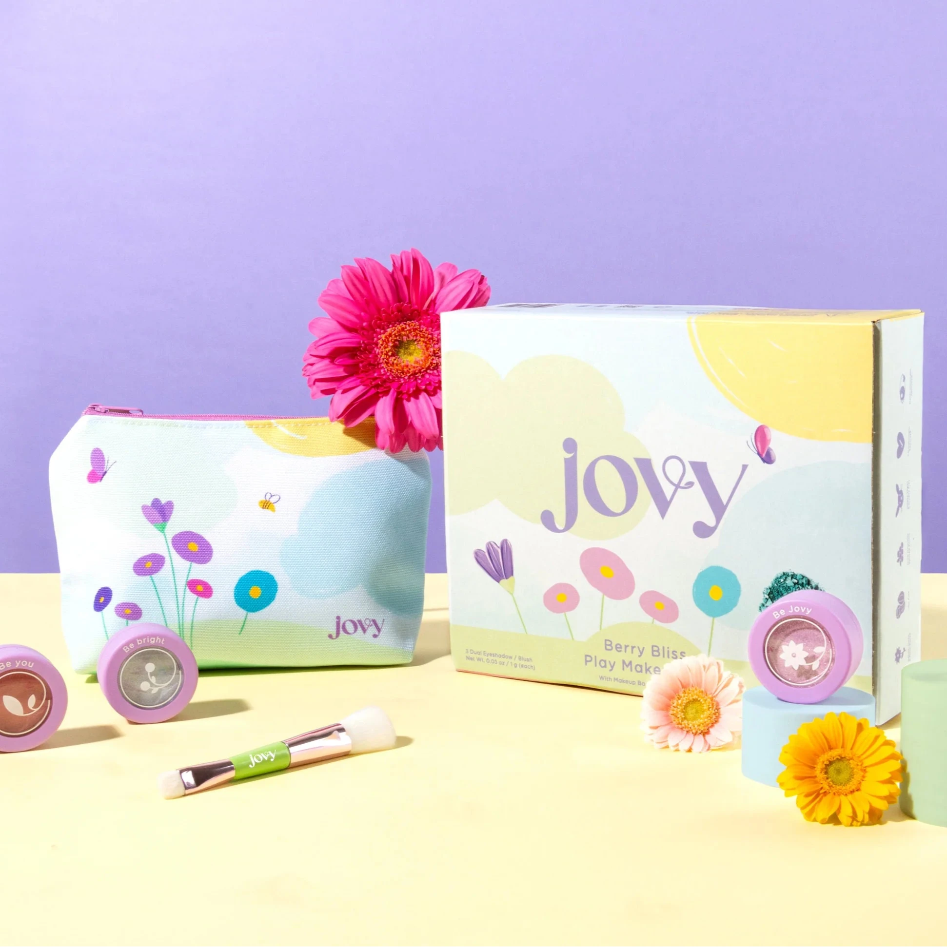 A collection of children's makeup products from Jovy, including colorful eyeshadows and a compact powder, displayed on a table with a floral theme.