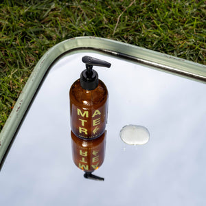 A bottle of mater hand soap on a mirror with grass in the background. 