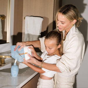 A woman helps a little girl refill the nemah body lotion in a bathroom setting. 