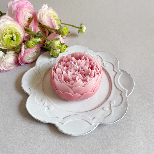A pink peony flower-shaped candle placed on a white decorative plate with fresh peony flowers in the background.