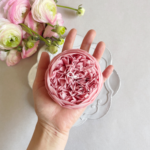 A person holding a pink peony candle above a bouquet and a white surface. 