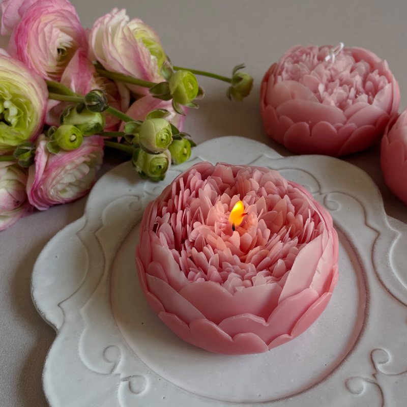 A pink peony flower-shaped candle placed on a white decorative plate with fresh peony flowers in the background.