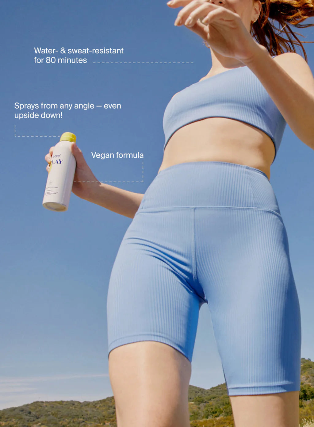 Person wearing blue athletic wear holding a spray bottle of PLAY mist against a clear blue sky.