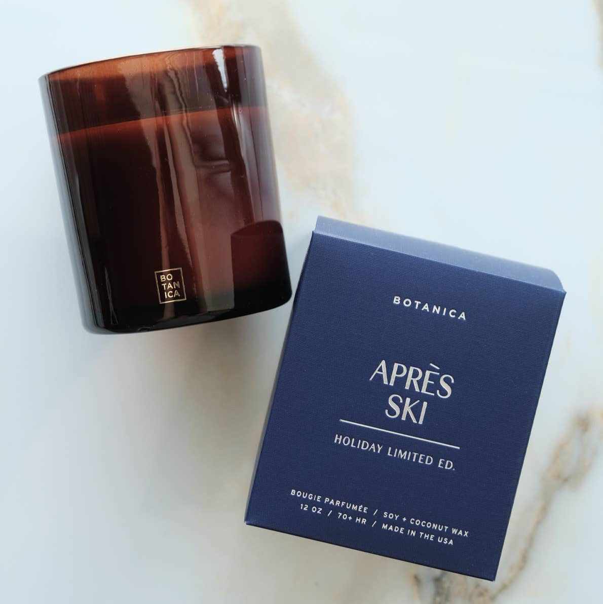 Candle with a lit wick next to its packaging on a dark background