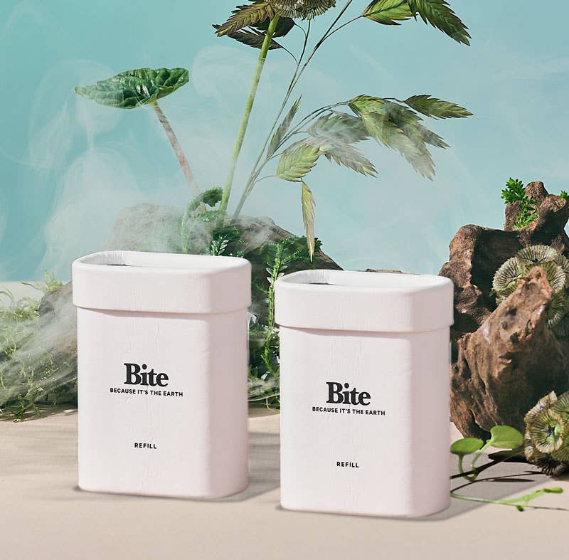The image features two cylindrical white deodorant containers with the brand name 'BITE' printed on them, placed against a backdrop of a misty forest with a large rock and greenery in the background. There is also a smaller container with a plant inside it, suggesting the product is related to deodorants.