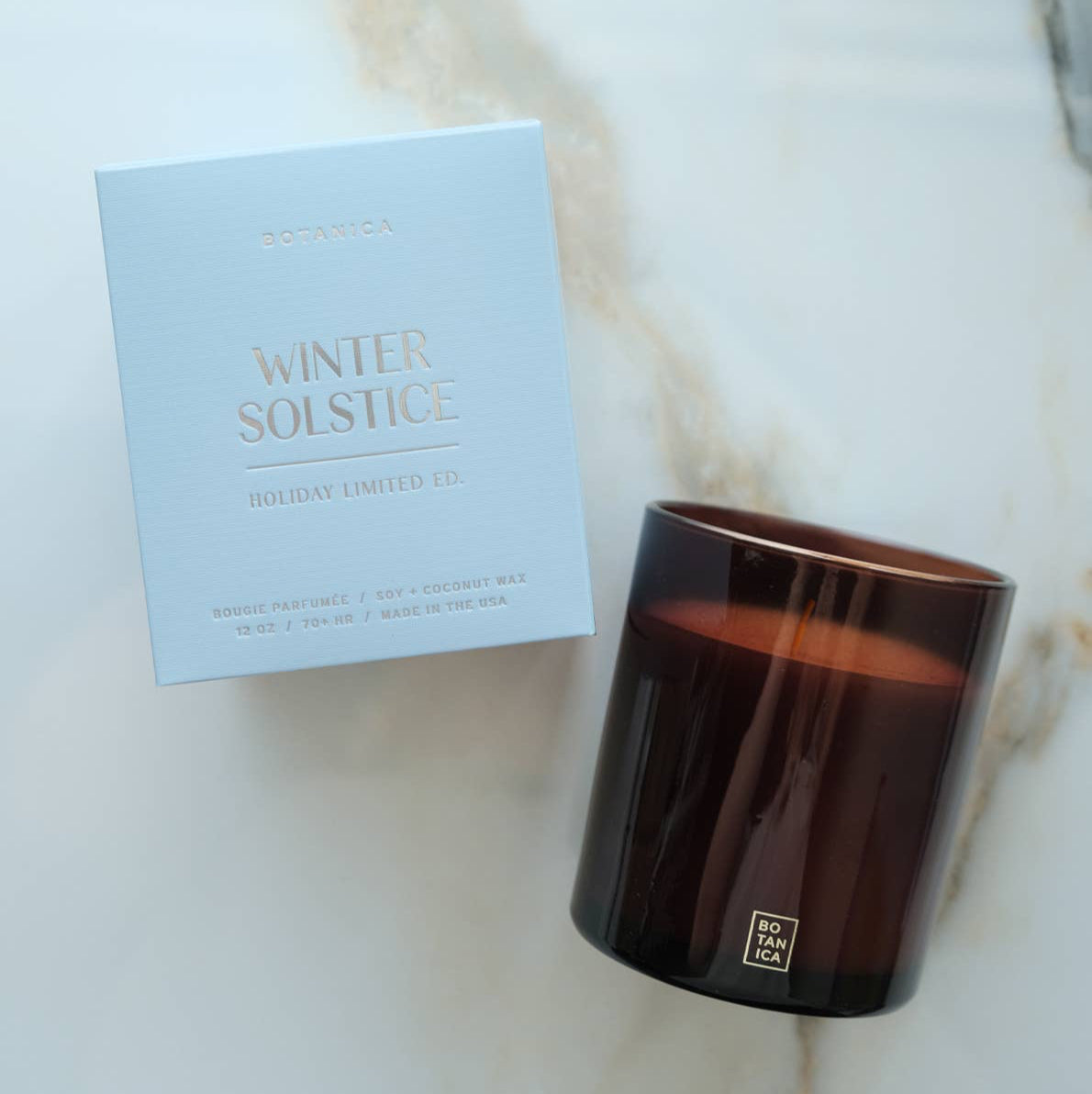 Lit candle in a dark container next to its packaging on a gray background
