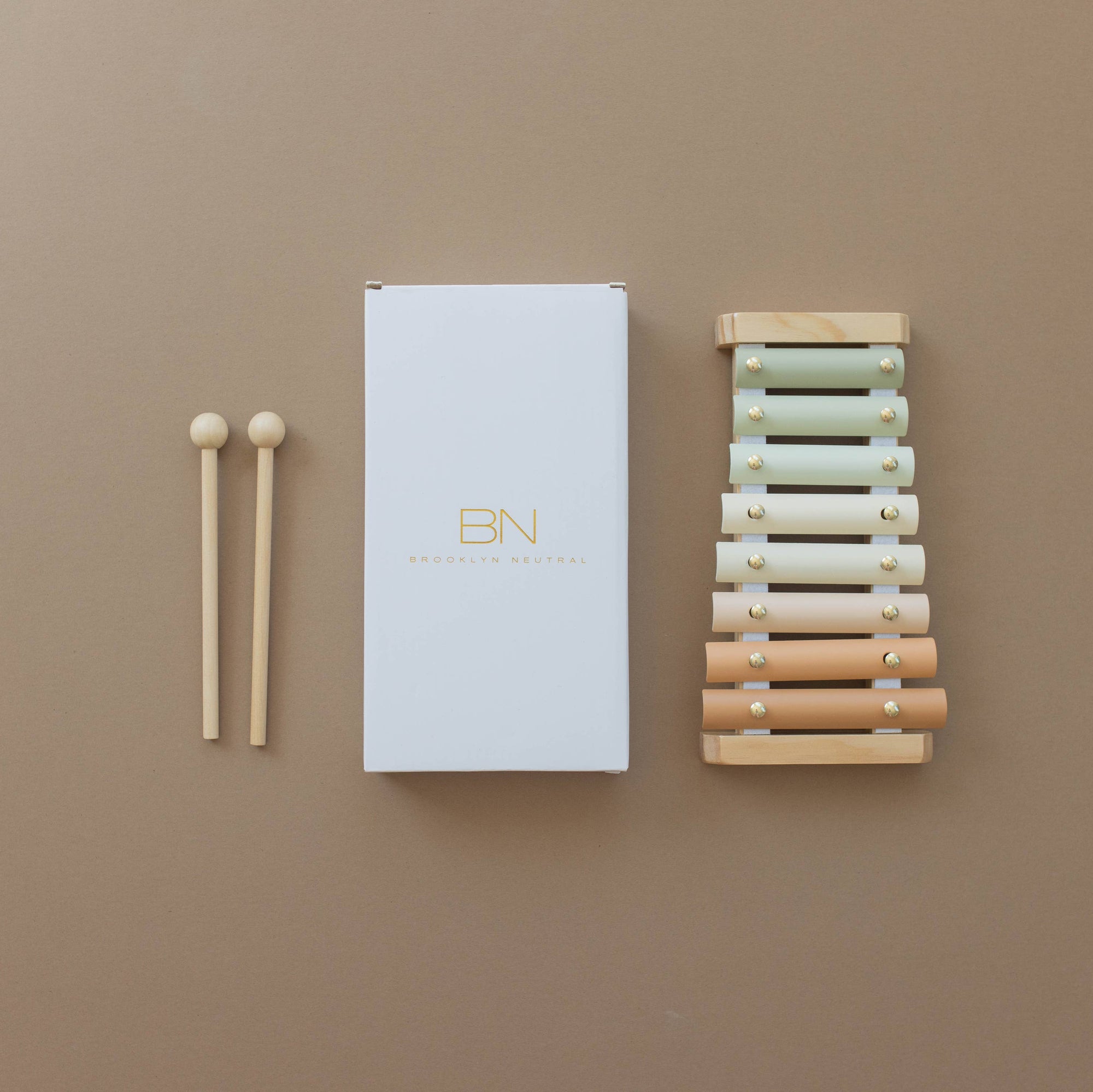 A neutral colored xylophone with wooden keys and metal accents, alongside its packaging box, placed on a white surface with green foliage in the background.